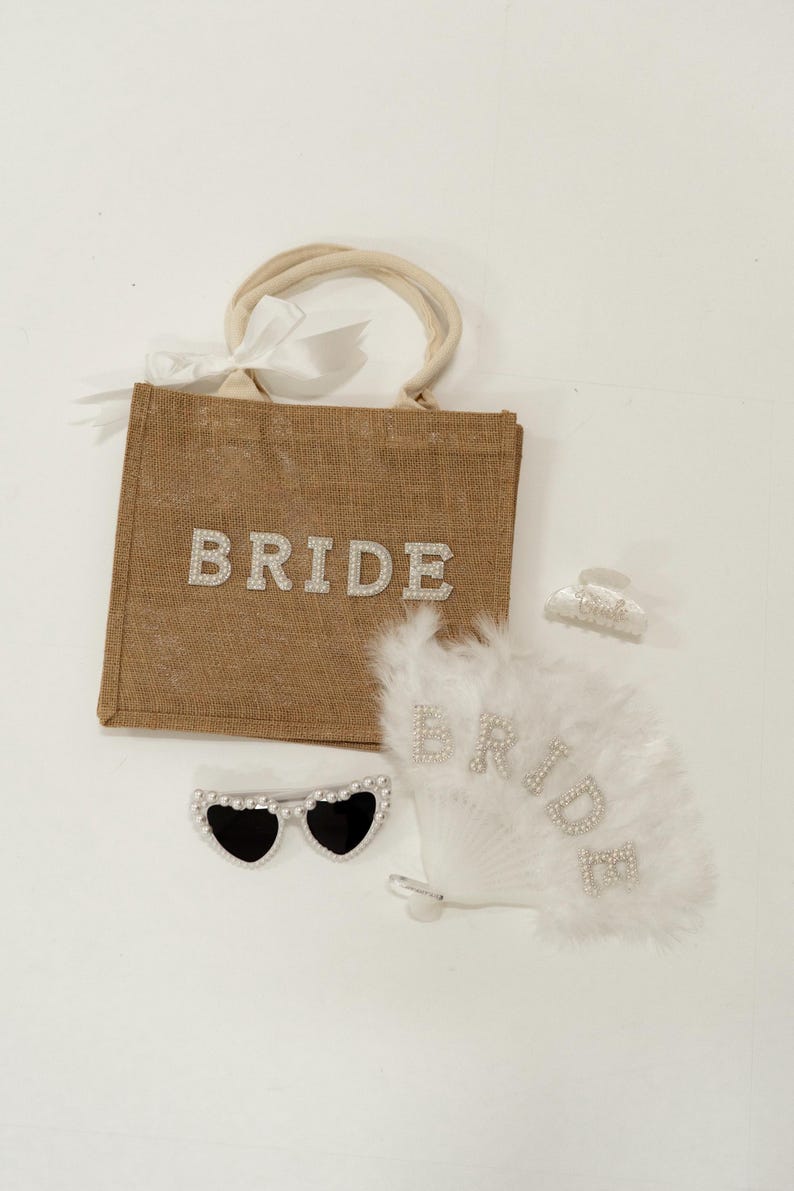 May include: A collection of bridal accessories, including a burlap tote bag with "BRIDE" in rhinestone letters, heart-shaped sunglasses, a feather fan, and a hair clip. The fan and bag also feature the word "BRIDE".