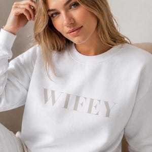 May include: White sweatshirt with the word "WIFEY" printed in a light tan color. The sweatshirt is a crew neck style and appears to be made of a soft material. The image shows a woman wearing the sweatshirt.