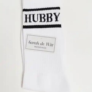 May include: White socks with the word "HUBBY" in black block letters across the top, framed by black stripes. A rectangular tag with the text "Sarah de Witt WEDDINGS" is attached to the sock.