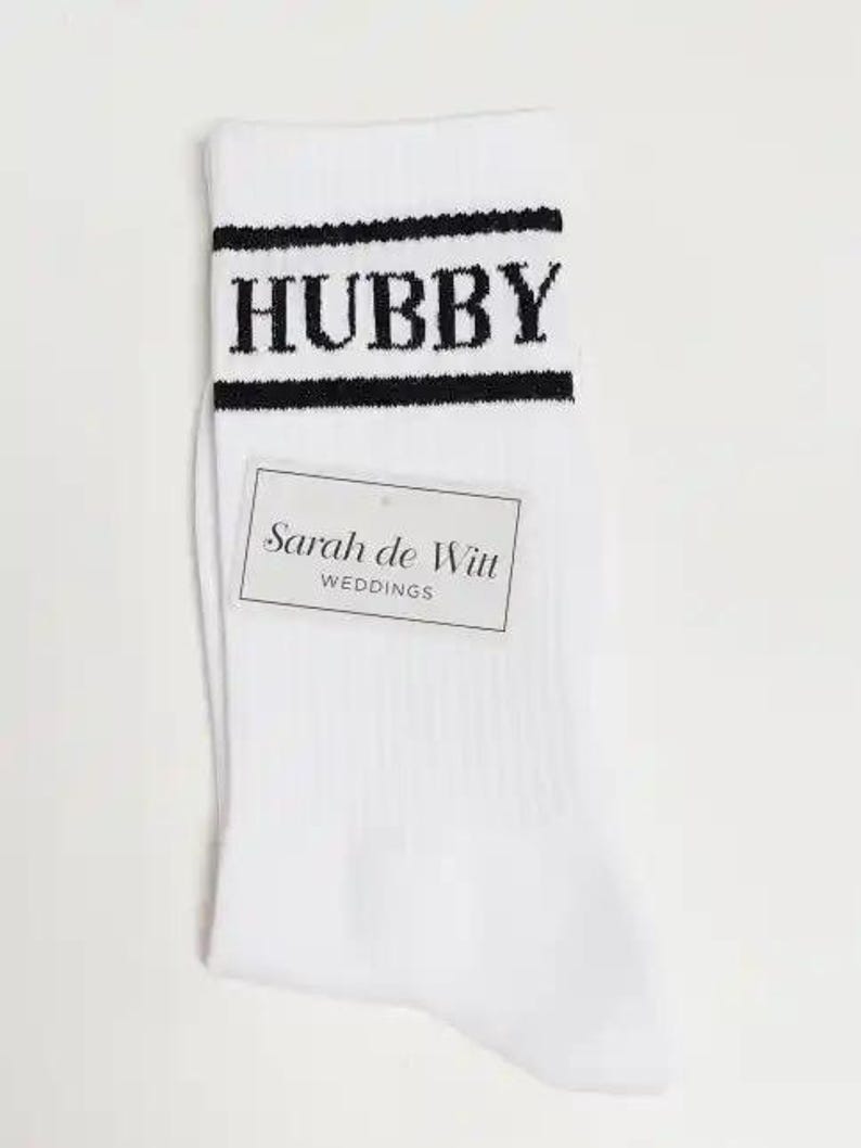 May include: White socks with the word "HUBBY" in black block letters across the top, framed by black stripes. A rectangular tag with the text "Sarah de Witt WEDDINGS" is attached to the sock.