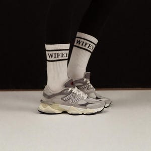 K&ouml;nnte beinhalten: Ein Paar graue und wei&szlig;e New Balance 9060 Sneaker mit wei&szlig;en Socken mit dem Wort "WIFEY" in schwarzen Buchstaben. Die Sneaker haben eine klobige Sohle und das New Balance Logo an der Seite.