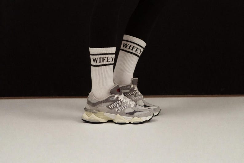 K&ouml;nnte beinhalten: Ein Paar graue und wei&szlig;e New Balance 9060 Sneaker mit wei&szlig;en Socken mit dem Wort "WIFEY" in schwarzen Buchstaben. Die Sneaker haben eine klobige Sohle und das New Balance Logo an der Seite.