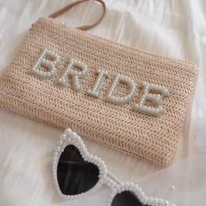 May include: A beige woven straw clutch bag featuring the word BRIDE in white pearl-encrusted letters, accompanied by heart-shaped sunglasses with white pearl-embellished frames and dark lenses, creating a coordinated set of accessories.