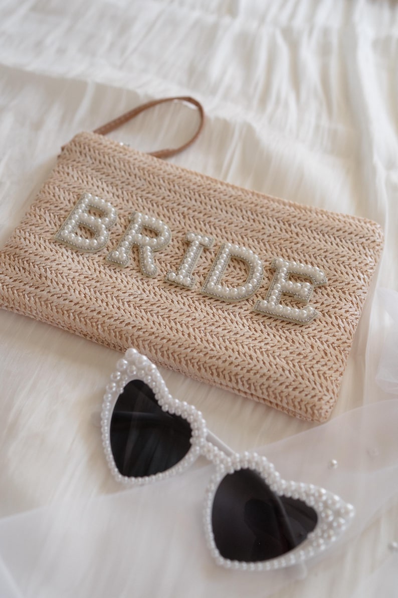 May include: A beige woven straw clutch bag featuring the word BRIDE in white pearl-encrusted letters, accompanied by heart-shaped sunglasses with white pearl-embellished frames and dark lenses, creating a coordinated set of accessories.
