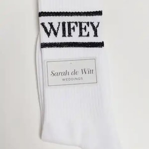 K&ouml;nnte beinhalten: Wei&szlig;e Socken mit dem Wort "WIFEY" in schwarzen Blockbuchstaben oben. Die Socken haben einen schwarzen Streifen &uuml;ber und unter dem Text. Ein rechteckiges Etikett lautet "Sarah de Witt WEDDINGS."