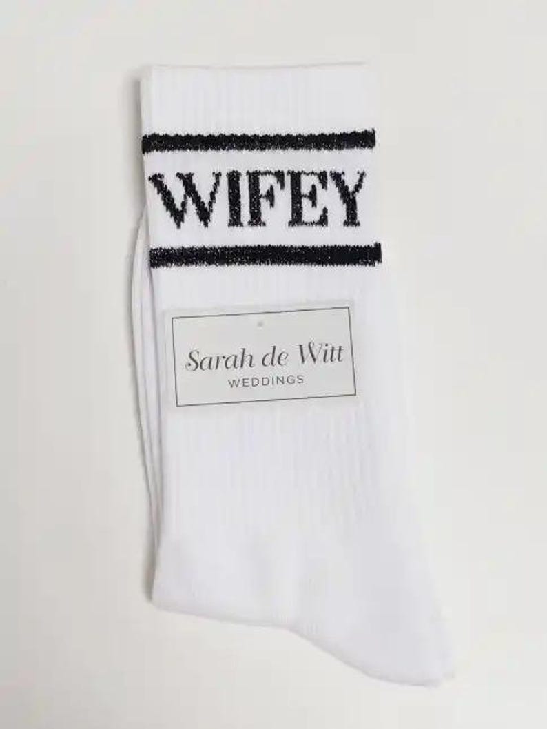 K&ouml;nnte beinhalten: Wei&szlig;e Socken mit dem Wort "WIFEY" in schwarzen Blockbuchstaben oben. Die Socken haben einen schwarzen Streifen &uuml;ber und unter dem Text. Ein rechteckiges Etikett lautet "Sarah de Witt WEDDINGS."