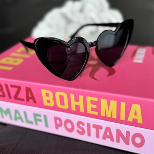 May include: Black heart-shaped sunglasses on top of a pink and white book with the text "Ibiza Bohemia Malfi Positano".