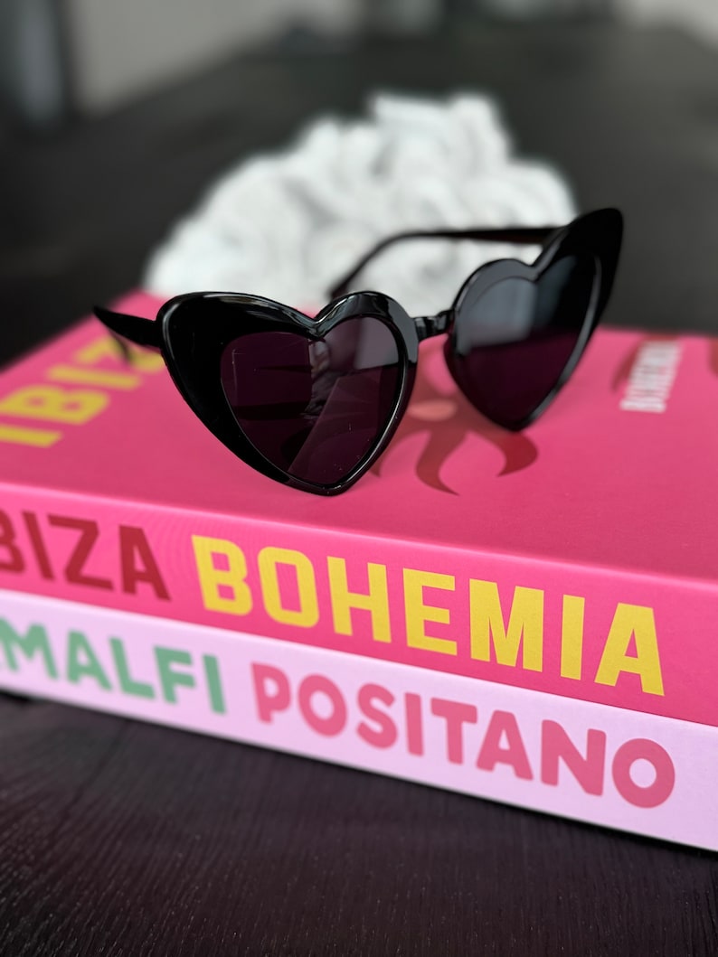 May include: Black heart-shaped sunglasses on top of a pink and white book with the text "Ibiza Bohemia Malfi Positano".