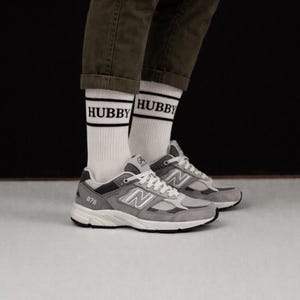 May include: A pair of gray and white New Balance 878 sneakers with white laces, paired with white socks featuring the word "HUBBY" in black block letters. The sneakers are set against a neutral background.