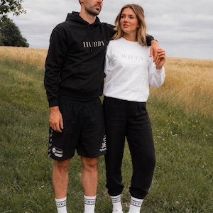 K&ouml;nnte beinhalten: Ein Paar in passender Kleidung im Freien. Der Mann tr&auml;gt einen schwarzen Hoodie mit dem Aufdruck "HUBBY" und schwarze Shorts. Die Frau tr&auml;gt ein wei&szlig;es Sweatshirt mit dem Aufdruck "WIFEY" und eine schwarze Hose. Beide tragen wei&szlig;e Turnschuhe und Socken.