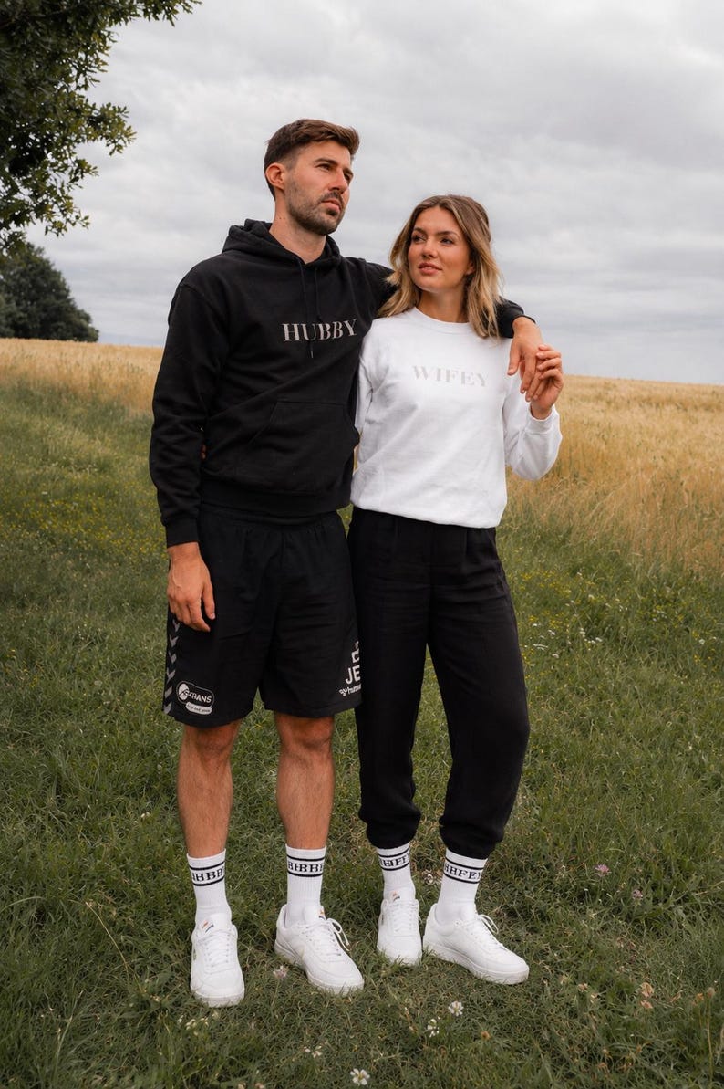 K&ouml;nnte beinhalten: Ein Paar in passender Kleidung im Freien. Der Mann tr&auml;gt einen schwarzen Hoodie mit dem Aufdruck "HUBBY" und schwarze Shorts. Die Frau tr&auml;gt ein wei&szlig;es Sweatshirt mit dem Aufdruck "WIFEY" und eine schwarze Hose. Beide tragen wei&szlig;e Turnschuhe und Socken.