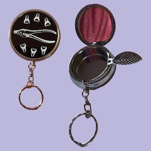 May include: Two pocket ashtrays with keychains. One is round with a black face, white teeth, and dental pliers. The other is open, silver, and has a leaf-shaped rest. Both have key rings.