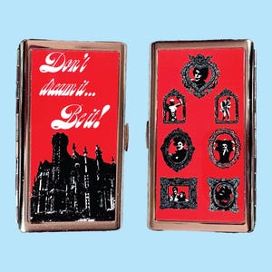 May include: A metal cigarette case with a red background. One side has the text "Don't dream it... Be it!" with a black silhouette of a building. The other side features framed black and white images.