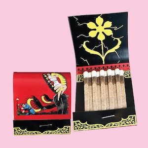 May include: A vintage matchbook with a black and red design. The front features a black and white illustration of a woman being swallowed by a plant. The back features a black background with a yellow flower and a black and white border.