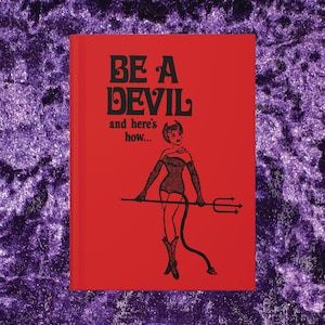 May include: A red hardcover book with black text that reads BE A DEVIL and here's how... The cover features a black line art illustration of a figure with horns, a tail, and a trident, wearing a textured bodysuit, gloves, and boots.