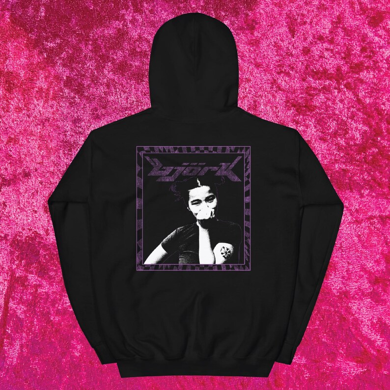 May include: Black hooded sweatshirt featuring a purple framed graphic of a person with the word "Bjork" above the image. The graphic is in black and white. The sweatshirt has a drawstring hood.