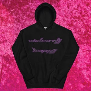 May include: Black hooded sweatshirt with the text "violently happy" in a purple, stylized font. The hoodie has a front pocket and a drawstring hood. The sweatshirt is displayed on a pink textured background.