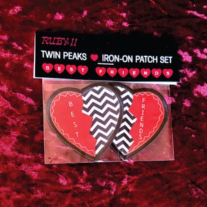 May include: A set of iron-on patches featuring a broken heart design. One half reads "BEST" in white letters on a red background, the other half reads "FRIENDS". The packaging reads "RUBY II TWIN PEAKS IRON-ON PATCH SET".