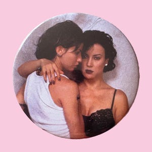 May include: Round button featuring a photograph of two women embracing. One woman wears a white tank top, the other a black lace top. The button has a pink background.