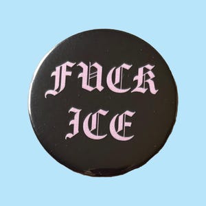 May include: A round, black button with the words "FUCK ICE" in pink gothic lettering. The button has a glossy finish and is set against a light blue background. The text is centered on the button.