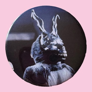 May include: A round button with a black and white image of a person in a menacing rabbit costume. The rabbit has large, pointed ears and a skeletal face. The button has a glossy finish and a dark background.
