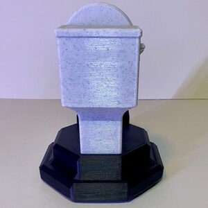 Puede incluir: Un portarrollos de papel higi&eacute;nico impreso en 3D, de color p&uacute;rpura claro con una textura moteada. El soporte tiene un cuerpo rectangular y una parte superior curvada, montado sobre una base geom&eacute;trica negra. La base tiene un dise&ntilde;o escalonado.
