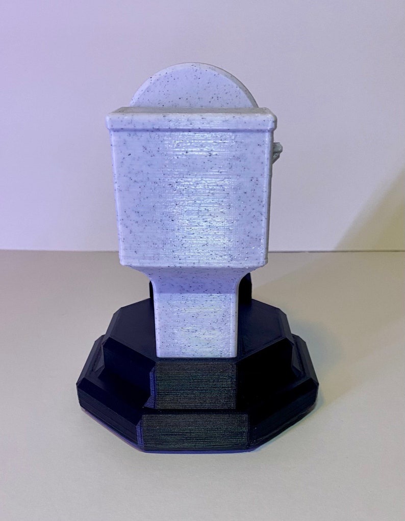 Puede incluir: Un portarrollos de papel higi&eacute;nico impreso en 3D, de color p&uacute;rpura claro con una textura moteada. El soporte tiene un cuerpo rectangular y una parte superior curvada, montado sobre una base geom&eacute;trica negra. La base tiene un dise&ntilde;o escalonado.
