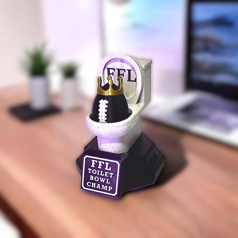 Puede incluir: Un trofeo novedoso que presenta un inodoro en miniatura con un objeto en forma de bal&oacute;n de f&uacute;tbol con una corona dorada. El inodoro tiene las letras "FFL" y la base dice "FFL TOILET BOWL CHAMP".