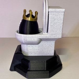 Puede incluir: Una figura de inodoro novedosa con un objeto negro en forma de huevo que lleva una corona dorada. El inodoro es blanco con motas negras y la base es negra. El asiento del inodoro est&aacute; levantado. Un art&iacute;culo decorativo humor&iacute;stico.