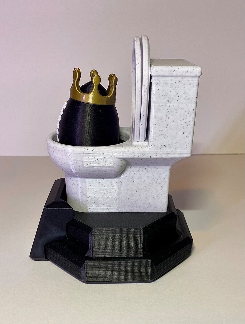 Puede incluir: Una figura de inodoro novedosa con un objeto negro en forma de huevo que lleva una corona dorada. El inodoro es blanco con motas negras y la base es negra. El asiento del inodoro est&aacute; levantado. Un art&iacute;culo decorativo humor&iacute;stico.