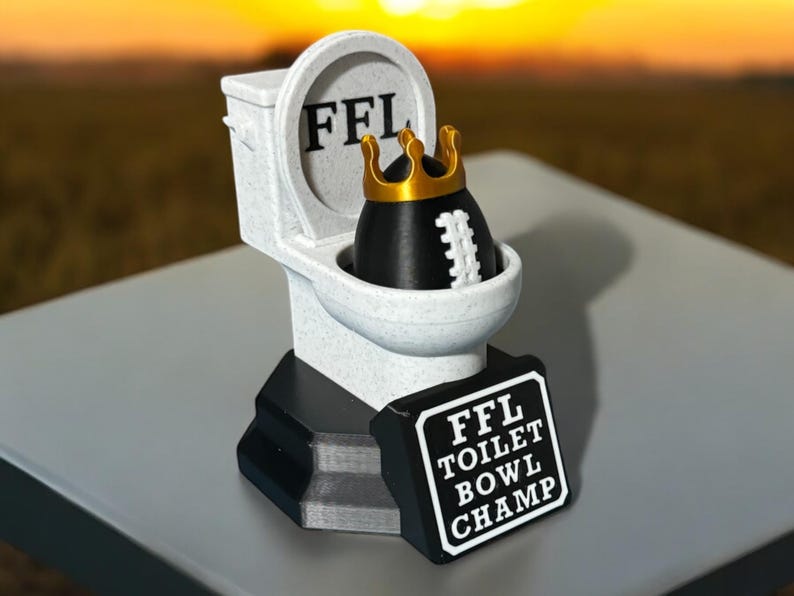 Puede incluir: Un trofeo novedoso que representa un inodoro blanco con las letras "FFL" en la tapa. Dentro del inodoro hay un bal&oacute;n de f&uacute;tbol americano negro con una corona dorada. Un letrero dice "FFL TOILET BOWL CHAMP".