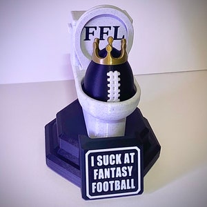 Puede incluir: Un trofeo novedoso con un bal&oacute;n de f&uacute;tbol dentro de un inodoro, coronado con una corona dorada. El inodoro tiene las letras "FFL" en la parte posterior. Un letrero en la base dice "I SUCK AT FANTASY FOOTBALL."