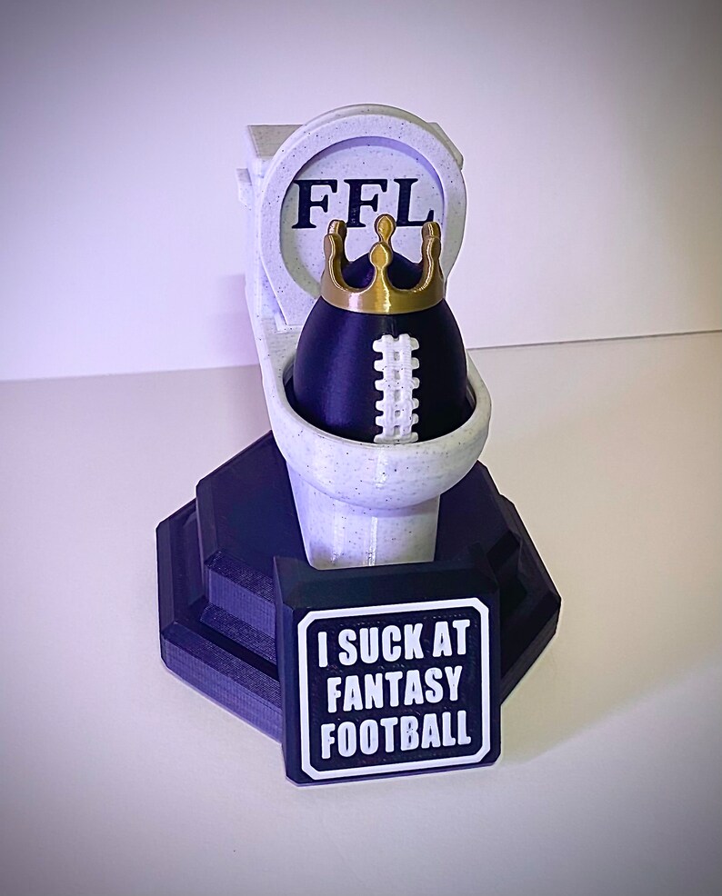 Puede incluir: Un trofeo novedoso con un bal&oacute;n de f&uacute;tbol dentro de un inodoro, coronado con una corona dorada. El inodoro tiene las letras "FFL" en la parte posterior. Un letrero en la base dice "I SUCK AT FANTASY FOOTBALL."