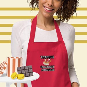Puede incluir: Un delantal rojo con el texto "Totsuki Culinary Academy" y una imagen de dibujos animados de un tazón de ramen. El delantal lo lleva una persona con cabello castaño y una camisa blanca.