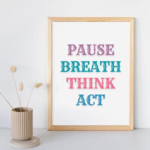 May include: A framed print with the words "PAUSE, BREATH, THINK, ACT" in colourful, stylised letters. The print is in a light wood frame and is displayed on a white surface. A beige vase with dried flowers is to the left of the frame.