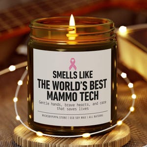 May include: A burning candle in a glass jar with a label stating "Smells Like The World's Best Mammo Tech." The candle is on a wooden coaster, with fairy lights around it. The label features a pink ribbon.