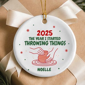 May include: A white ceramic Christmas ornament with the text "2025 The Year I Started Throwing Things" in red and green. The ornament features a pottery wheel design and the name "Noelle". The ornament is hanging on a gold string, and is placed on a wrapped gift.