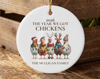 May include: A round white ceramic ornament hanging from a gold string. It features five chickens wearing holiday hats and scarves with colorful lights. Text reads: 2026 THE YEAR WE GOT CHICKENS and THE MULLIGAN FAMILY in dark brown lettering.
