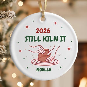 May include: A round white ceramic ornament hanging from a gold ribbon. The design features red text reading 2026, green text reading STILL KILN IT, and the name NOELLE in green. A red illustration shows hands shaping a clay pot on a pottery wheel.