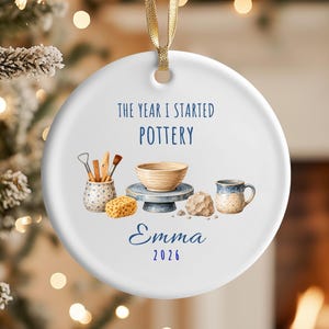 May include: A round white ceramic ornament hanging from a gold ribbon. The design features pottery tools, a bowl on a wheel, a sponge, and a mug. Text reads: THE YEAR I STARTED POTTERY, Emma, and 2026. The illustration is centered on the ornament.