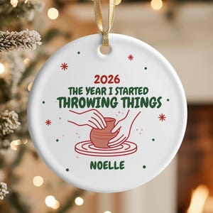 May include: A round white ceramic ornament with a gold ribbon hanger. The design features red and green text reading 2026 THE YEAR I STARTED THROWING THINGS and NOELLE, with a red graphic of hands shaping a clay pot on a pottery wheel.