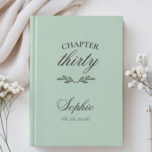 May include: A light green hardback book featuring dark grey text that reads CHAPTER thirty, followed by a decorative leaf flourish, the name Sophie, and the date 06.26.2026 printed in a combination of serif and elegant script fonts.