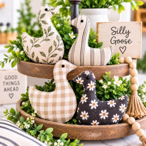 May include: Four fabric goose ornaments with black bead eyes. One has a green leaf print, one has vertical stripes, one has a beige gingham pattern, and one has a black daisy print. A wooden sign reads Silly Goose with a heart symbol.