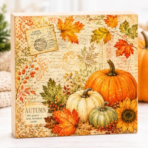 May include: A rectangular wooden block sign featuring a vintage-style print of orange and green pumpkins, a sunflower, and autumn leaves. The background includes script text, a postal stamp, and the words AUTUMN the year's last, loveliest smile.