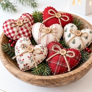 May include: A collection of six fabric heart ornaments nestled in a wooden bowl with pine sprigs. Each heart features a unique pattern including red gingham, solid red, cream with red berries, cream with stripes, and cream with green trees.