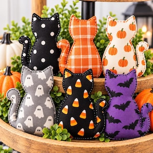 Peut inclure: Six ornements en forme de chat en tissu avec divers motifs : noir à pois blancs, écossais orange, blanc à citrouilles orange, gris à fantômes blancs, noir à bonbons maïs et violet à chauves-souris noires, disposés sur un plateau à étages.