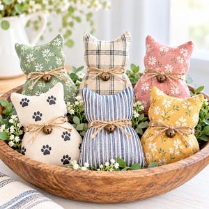May include: Five stuffed fabric cat-shaped ornaments in a wooden bowl. Each cat features a different pattern: green floral, cream plaid, pink floral, cream with black paw prints, blue striped, and yellow floral. Each has a jute twine bow and a small bell.