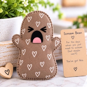 May include: A brown fabric plush toy shaped like a ghost with a white heart pattern, black stitched eyes, and an open mouth with a pink tongue. Beside it is a rectangular tan card with the text Scream Bean and an encouraging message.
