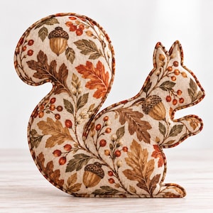 May include: A plush squirrel-shaped decorative pillow featuring a cream-colored fabric base with a printed pattern of brown oak leaves, acorns, and small red berries. The edges are finished with a matching brown fabric piping trim.