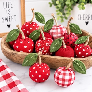 May include: A collection of fabric cherry ornaments in a wooden bowl. Each cherry features a red fabric body with various patterns including white polka dots, hearts, flowers, and red gingham, topped with a jute stem and a green felt leaf.